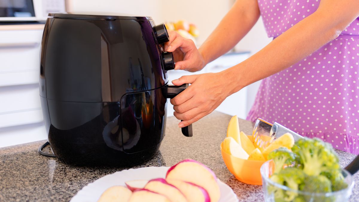 Pessoa utilizando uma air fryer preta em cozinha moderna com frutas e legumes na bancada — eletrodoméstico que economiza tempo e dinheiro