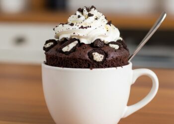 Bolo de Oreo na caneca em uma xícara branca, decorado com chantilly e biscoitos Oreo triturados, em uma mesa de madeira.