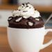 Bolo de Oreo na caneca em uma xícara branca, decorado com chantilly e biscoitos Oreo triturados, em uma mesa de madeira.