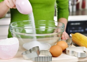 Pessoa medindo açúcar com xícara medidora rosa em tigela de vidro na cozinha, acompanhada de ovos, farinha e cortadores de biscoito — ilustrando como medir ingredientes corretamente.