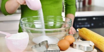 Pessoa medindo açúcar com xícara medidora rosa em tigela de vidro na cozinha, acompanhada de ovos, farinha e cortadores de biscoito — ilustrando como medir ingredientes corretamente.