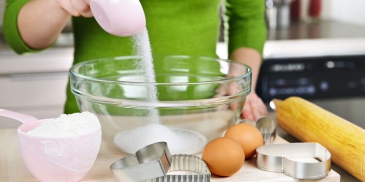 Pessoa medindo açúcar com xícara medidora rosa em tigela de vidro na cozinha, acompanhada de ovos, farinha e cortadores de biscoito — ilustrando como medir ingredientes corretamente.